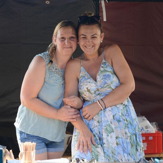 Zwei Frauen stehen eng beieinander, lächeln und posieren für ein Foto vor einem Zelt. Eine trägt ein ärmelloses Blumenkleid, die andere ein hellblaues Oberteil und Jeansshorts.