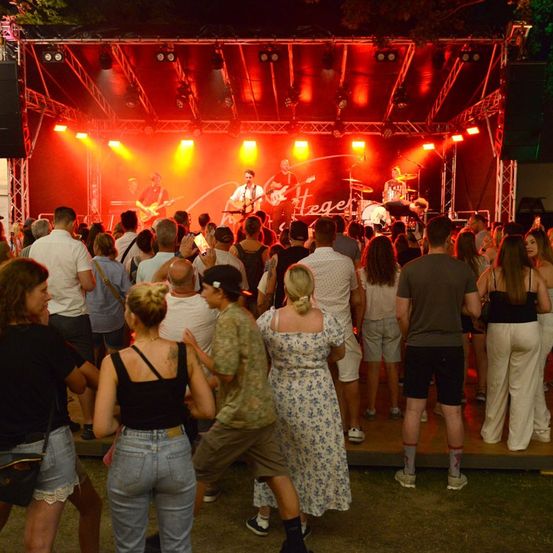 Eine Band spielt auf einer Bühne mit roter und gelber Beleuchtung, umgeben von einer Menge tanzender Menschen, die die Musik bei Nacht genießen.