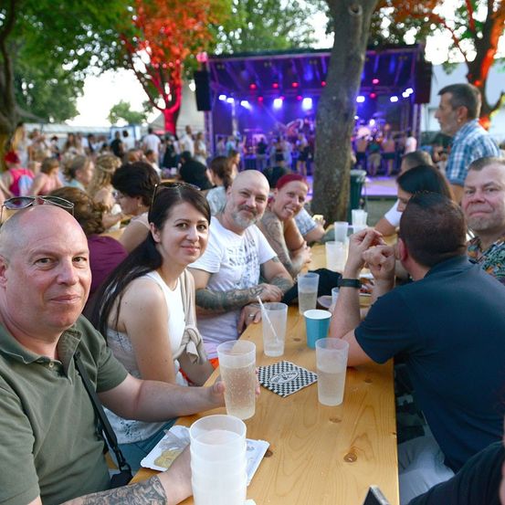 Eine Gruppe von Menschen sitzt an einem langen Tisch unter einem Baum, lächelt und genießt ein Live-Musik-Event. Der Tisch ist mit Gläsern, Tellern und Servietten gedeckt. Im Hintergrund eine Bühne mit lila Beleuchtung und eine Menschenmenge.