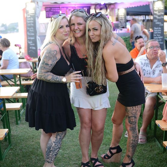 Drei Frauen mit Tattoos stehen auf einer Rasenfläche, wahrscheinlich bei einem Festival. Sie lächeln und posieren für ein Foto. Hinter ihnen sitzen Leute auf Bänken und Tischen, und ein Mann isst. Es gibt auch Essensstände und Zelte in der Gegend.