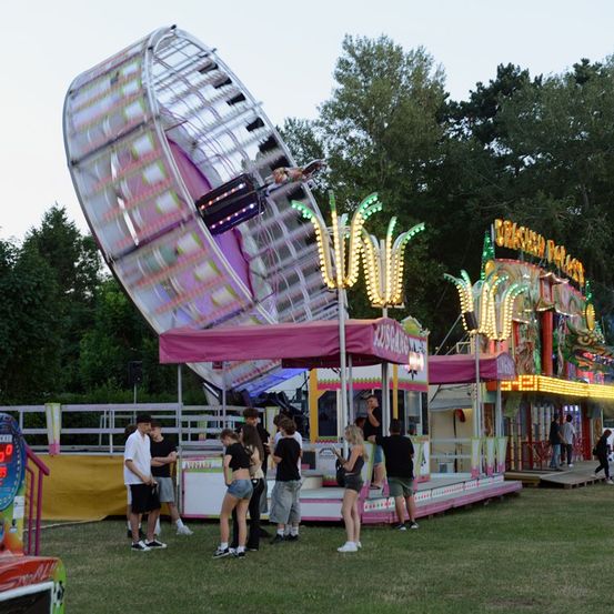 Eine Gruppe von Menschen steht um eine bunte Vergnügungsfahrt mit einem großen Riesenrad herum, umgeben von anderen Fahrgeschäften und Bäumen.