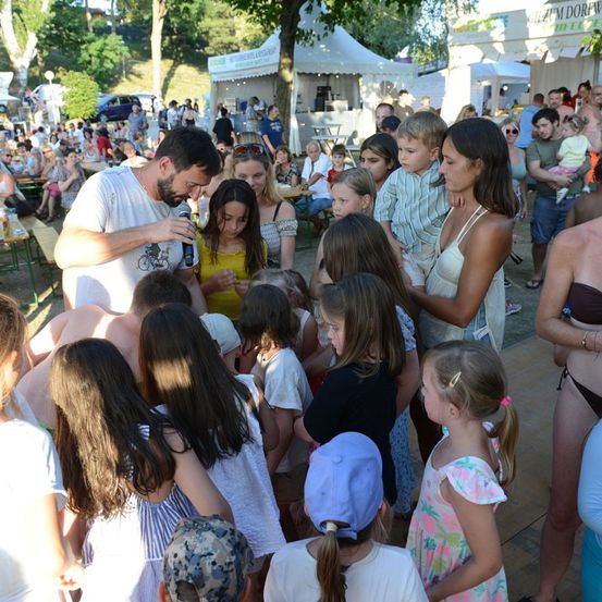 Eine Gruppe von Menschen, einschließlich Kindern, versammelt sich in einer Outdoor-Umgebung. Ein Mann hält ein Mikrofon und scheint zu sprechen. Die Menge ist vielfältig, mit verschiedenen Altersgruppen und Geschlechtern.