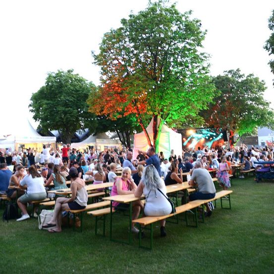 Eine große Menschenmenge ist für eine Veranstaltung im Freien versammelt. Sie sitzen an Picknicktischen unter Bäumen mit bunten Lichtern. Im Hintergrund befindet sich eine Bühne mit einer großen Leinwand und einem Zelt.
