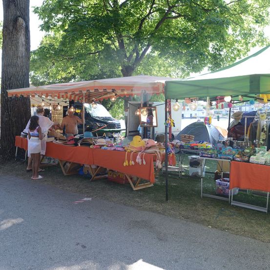 Ein Straßenmarkt mit zwei Zelten, einem grünen und einem orangefarbenen, die verschiedene Artikel im Schatten von Bäumen präsentieren. Menschen stöbern und ein Mann steht hinter einem Tisch.