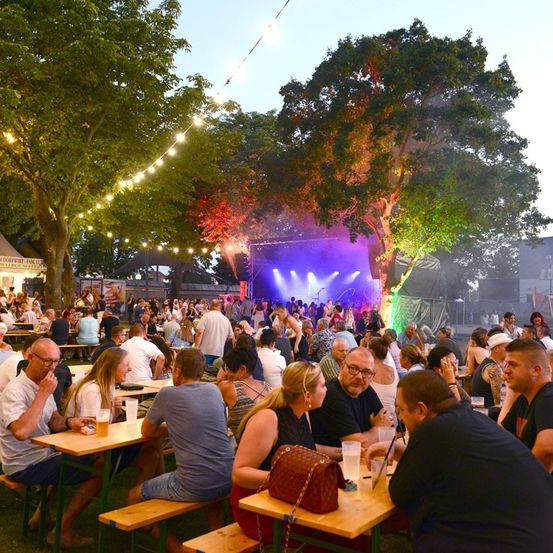 Ein lebhaftes Open-Air-Event mit Menschen an Tischen unter Bäumen, einer Bühne mit bunten Lichtern und einer lebhaften Atmosphäre.