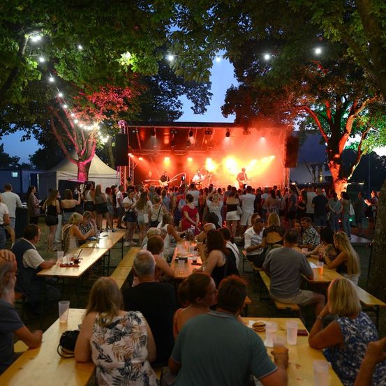 Ein nächtliches Open-Air-Konzert mit einer Band auf der Bühne, beleuchtet von roten und gelben Lichtern, mit einer Menschenmenge, die an Tischen unter Bäumen sitzt.