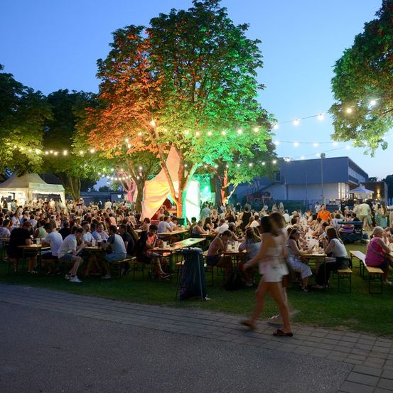 Eine Outdoor-Veranstaltung bei Dämmerung mit vielen Menschen, die an Tischen unter Bäumen und Zelten sitzen, beleuchtet von bunten Lichtern. Eine Frau geht an den Tischen vorbei.