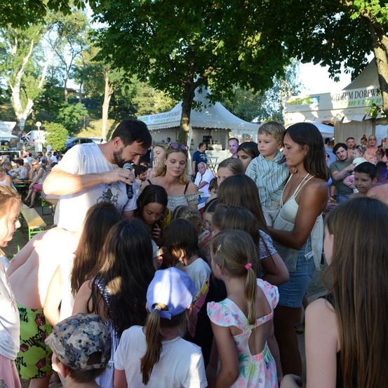 Ein Mann mit Mikrofon wird von einer Gruppe von Kindern und Erwachsenen in einem Park umringt. Der Mann spricht zu den Kindern, und sie hören aufmerksam zu. Der Park ist voller Menschen, und es gibt Bäume und Zelte im Hintergrund.