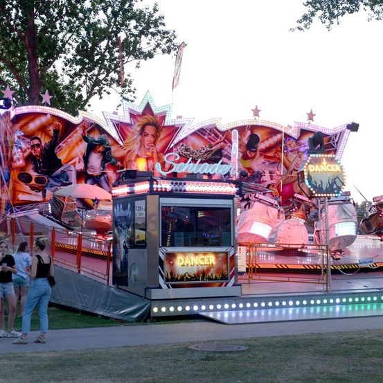 Ein leuchtendes Jahrmarkt-Karussell namens 'Schlander' mit Neonlichtern und Bildern tanzender Menschen, im Freien mit Leuten in der Nähe.