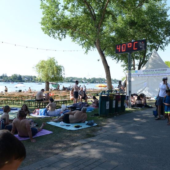 Ein Park am See mit sonnenbadenden Menschen, einem digitalen Thermometer mit 40°C und einem Zelt im Hintergrund.