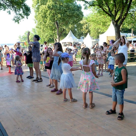 Eine Gruppe von Kindern und Erwachsenen tanzt auf einer Holzbühne bei einer Outdoor-Veranstaltung, umgeben von Bäumen und Zelten.