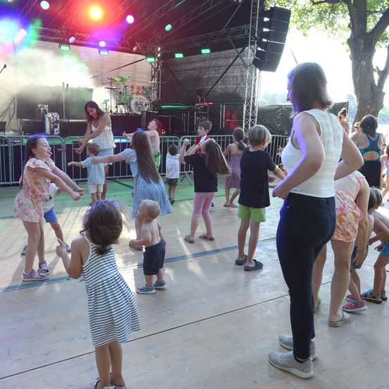 Eine Gruppe von Kindern und Erwachsenen tanzt auf einer Bühne unter bunten Lichtern, während eine Band im Hintergrund auftritt.