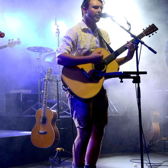 Ein Mann auf der Bühne spielt eine Gitarre und singt in ein Mikrofon, mit anderen Instrumenten und Bühnenlichtern im Hintergrund.