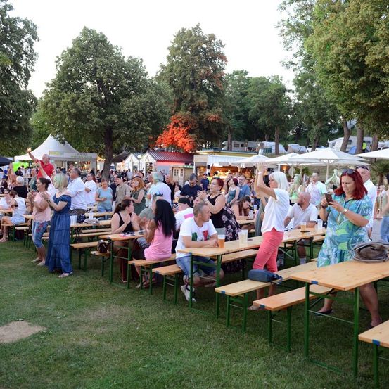 Eine große Menschenmenge versammelt sich in einem Park im Freien, sitzt an Picknicktischen unter weißen Sonnenschirmen. Einige klatschen und tanzen, während andere essen und trinken. Der Park ist von Bäumen umgeben und im Hintergrund befinden sich Zelte und Stände.