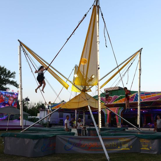 Eine Gruppe von Menschen genießt eine Schaukelfahrt auf einem Jahrmarkt, wobei eine Person in der Luft ist und andere am Boden warten. Die Fahrt ist hell beleuchtet und von bunten Dekorationen umgeben.