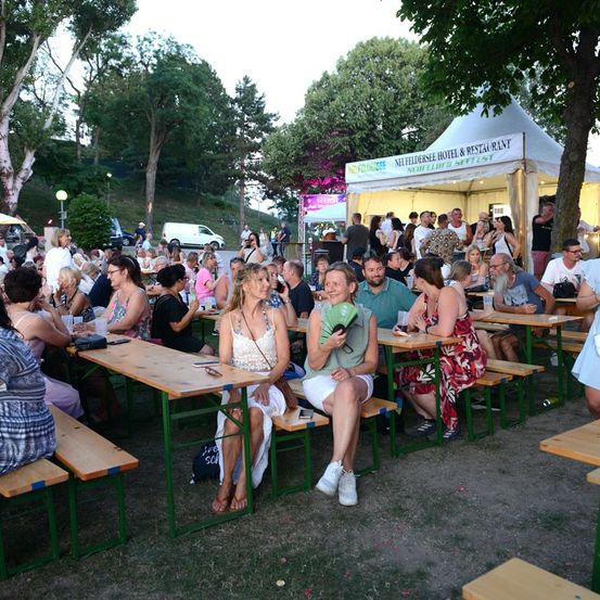 Eine Gruppe von Menschen sitzt an Tischen in einem Außenbereich, wahrscheinlich bei einem Festival. Einige tragen Brillen und einige halten Gläser. Ein Zelt mit einem Schild ist in der Ferne.