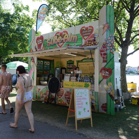 Ein Crepe-Stand mit einer bunten Wandmalerei und einem Menübrett draußen. Eine Frau in einem Bikini geht daran vorbei.