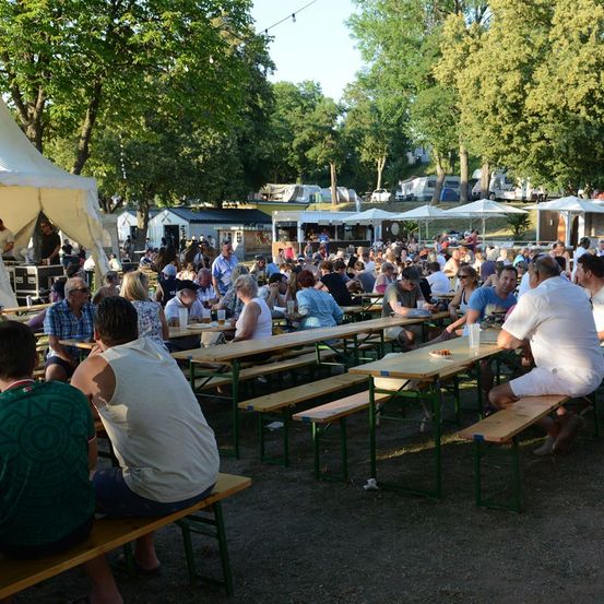 Eine große Gruppe von Menschen sitzt an Picknicktischen in einem Außenbereich mit Bäumen und Zelten im Hintergrund. Einige unterhalten sich, während andere ihre Mahlzeiten genießen.