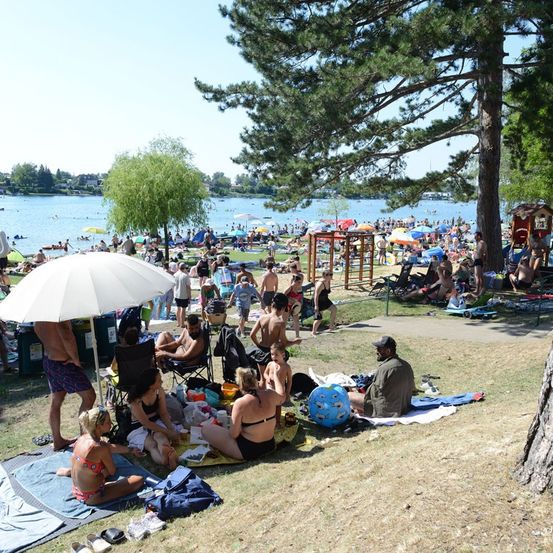 Ein überfüllter Strand mit Menschen, die sich sonnen, schwimmen und unter Sonnenschirmen in der Nähe eines Sees entspannen.