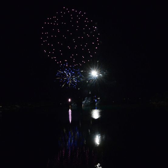 Ein nächtliches Feuerwerk mit einem Ausbruch von blauen und rosafarbenen Feuerwerken am Himmel, das sich im Wasser darunter spiegelt.