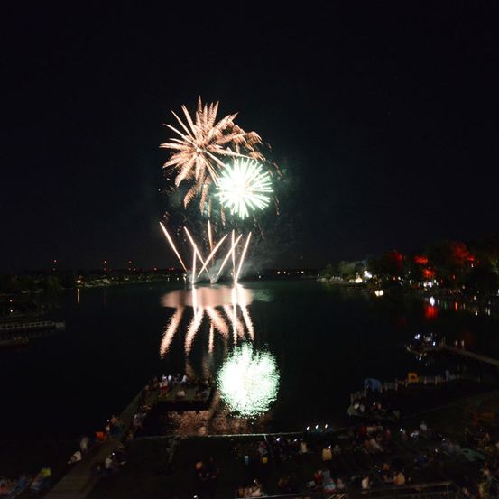 Ein nächtliches Feuerwerk über einem See mit Zuschauern am Ufer und Reflexionen im Wasser.