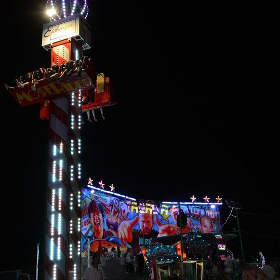 Ein nächtliches Achterbahn-Fahrgeschäft mit einem großen Schild, das 'Moser Rides' und bunte Lichter zeigt. Menschen sitzen auf der Fahrt, und eine große Leinwand zeigt Bilder von Menschen.
