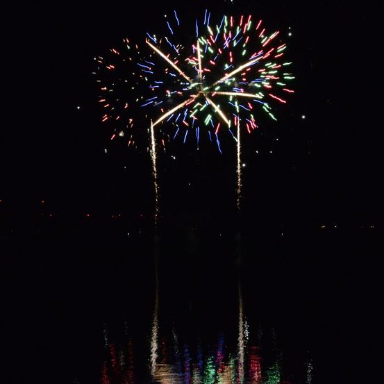 Ein leuchtendes Feuerwerk explodiert in der Nacht über einer ruhigen Wasserfläche und spiegelt sich farbig auf der Oberfläche wider.