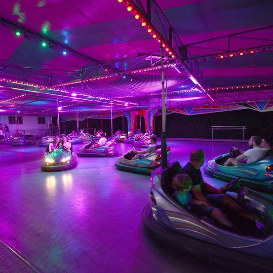 Eine Gruppe von Menschen genießt Kollisionsautos in einem hell beleuchteten Indoor-Vergnügungspark mit lila Beleuchtung.