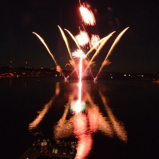 Ein nächtliches Feuerwerk über einem See mit Zuschauern auf einem Steg, das sich im Wasser spiegelt.