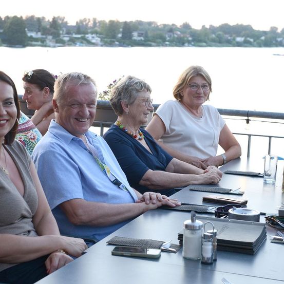 Eine Gruppe von Menschen, darunter eine ältere Frau in einem blauen Shirt, sitzt um einen Tisch auf einem Balkon, lächelt und schaut in die Kamera. Sie sind umgeben von einer malerischen Aussicht auf einen See und Bäume.