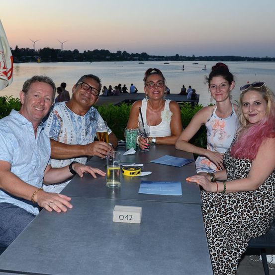 Fünf Personen sitzen um einen Tisch herum, lächeln und posieren für ein Foto. Sie halten Getränke in den Händen und im Hintergrund ist ein See und ein Sonnenuntergang zu sehen.