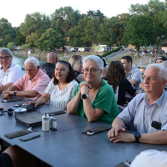Eine Gruppe älterer Erwachsener sitzt an einem Tisch in einem Park, mit einem See und Bäumen im Hintergrund.