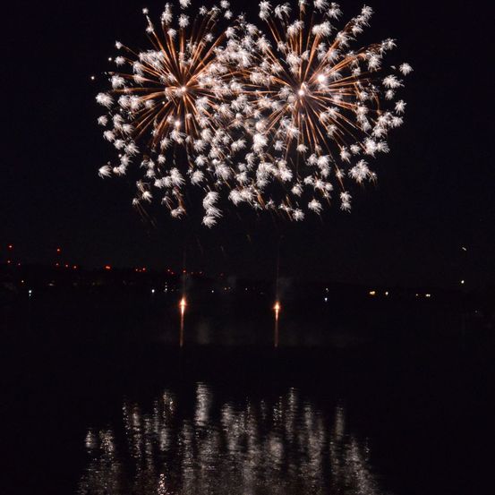 Ein nächtliches Feuerwerk explodiert am Himmel über einem Gewässer und spiegelt sich auf der Oberfläche darunter.