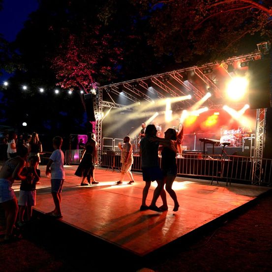 Ein nächtliches Open-Air-Konzert mit Bühne, bunten Lichtern und einer Gruppe tanzender Menschen.
