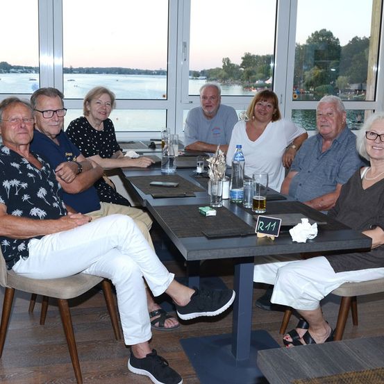 Eine Gruppe älterer Menschen sitzt in einem Restaurant mit Blick auf einen See und Berge.