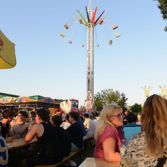 Eine Gruppe von Menschen sitzt an Tischen in einem Außenbereich mit einem Riesenrad im Hintergrund. Das Riesenrad ist bunt mit mehreren Kabinen. Der Himmel ist klar und blau.