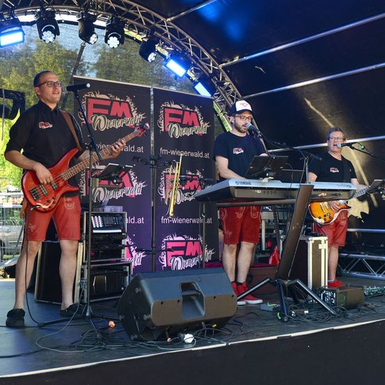 Drei Männer spielen auf einer Bühne mit Musikinstrumenten. Der Mann auf der linken Seite spielt eine rote Bassgitarre, der Mann in der Mitte spielt ein Keyboard und der Mann auf der rechten Seite spielt eine orange Gitarre. Sie stehen unter einem Zelt mit einem Banner, das FM Wienerwald liest.