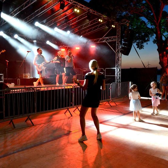 Eine Band spielt auf der Bühne bei Dämmerung mit roten und weißen Lichtern, während eine Frau und zwei Mädchen vor der Bühne tanzen.