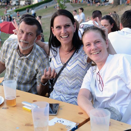 Drei Menschen sitzen an einem Tisch in einer Außenumgebung, lächeln und posieren für ein Foto. Der Mann auf der linken Seite hat einen Bart und macht ein Friedenszeichen. Die Frau in der Mitte hat eine Halskette und eine Tasche. Die Frau auf der rechten Seite trägt eine Brille und ein weißes Hemd. Auf dem Tisch liegen Gläser, ein Handy und ein Buch. Im Hintergrund sitzen Menschen auf dem Rasen.