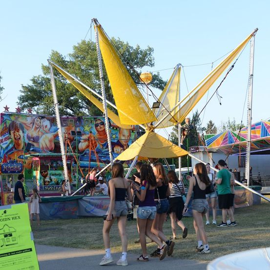 Eine Gruppe von Menschen geht an einem großen gelben Schaukelspielplatz auf einem Freiluftfest vorbei, mit bunten Buden und Fahrgeschäften im Hintergrund.
