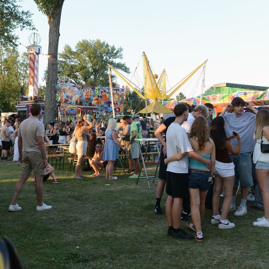 Eine Gruppe von Menschen versammelt sich auf einem Freiluftfest, mit Fahrgeschäften und Attraktionen im Hintergrund. Einige stehen, andere sitzen an Tischen.