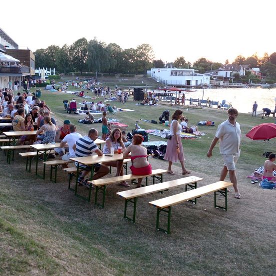 Eine große Gruppe von Menschen versammelt sich im Freien in der Nähe eines Sees, einige sitzen an Picknicktischen, andere liegen auf der Wiese. Die Sonne geht im Hintergrund unter.