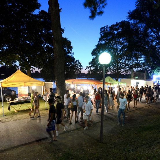 Ein nächtlicher Outdoor-Markt mit Zelten, vorbeigehenden Menschen und einer Straßenlaterne. Der Markt ist von Bäumen und einem Gewässer umgeben.