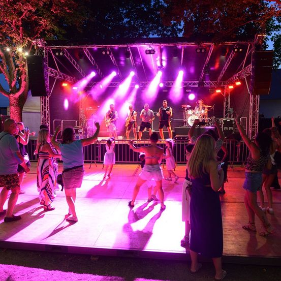 Ein nächtliches Open-Air-Konzert mit einer Band auf der Bühne unter lila Lichtern, vor der Menschen tanzen.