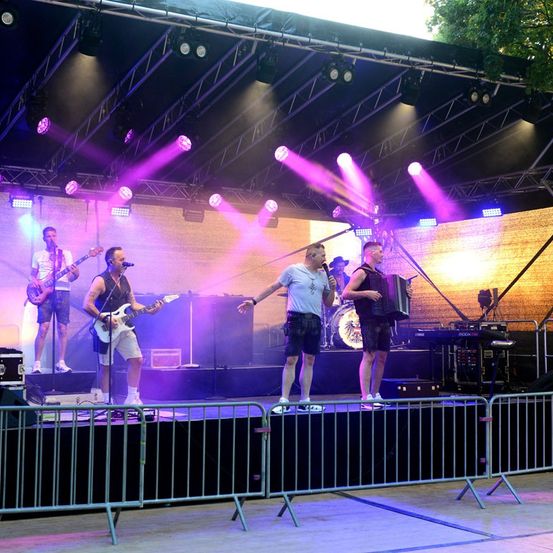Eine Band spielt auf der Bühne mit lila und blauer Beleuchtung. Ein Mitglied spielt Gitarre, ein anderes Akkordeon und der Schlagzeuger steht hinter einem Schlagzeug.