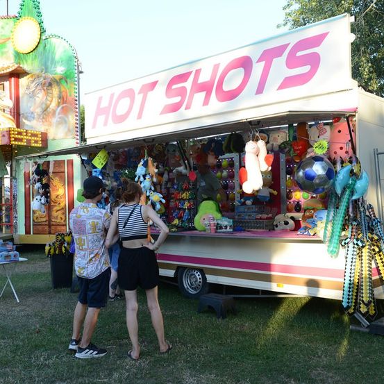 Ein Mann und eine Frau stehen vor einem 'Hot Shots'-Karnevalspiel, mit Kuscheltieren, die an den Haken des Spiels hängen.