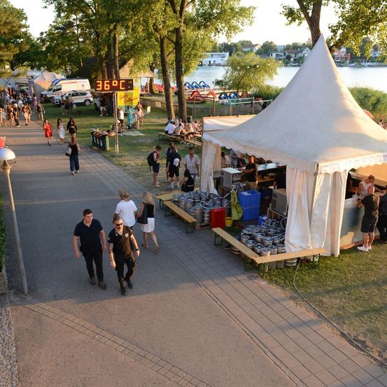 Ein Outdoor-Event mit Menschen, die gehen, einem großen Zelt und einem digitalen Temperaturanzeiger, der 36 Grad Celsius anzeigt.
