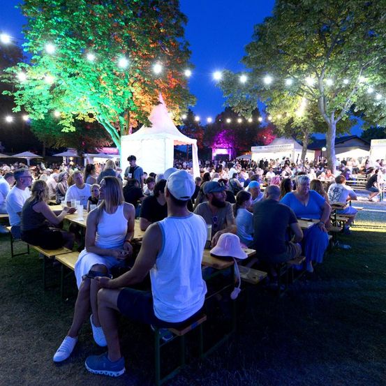 Eine Gruppe von Menschen versammelt sich in einem Außenbereich, möglicherweise einem Park, für eine Veranstaltung bei Nacht. Sie sitzen auf Bänken um Tische herum, und einige stehen. Der Bereich ist von bunten Lichtern beleuchtet, und es gibt Bäume und Zelte im Bereich.