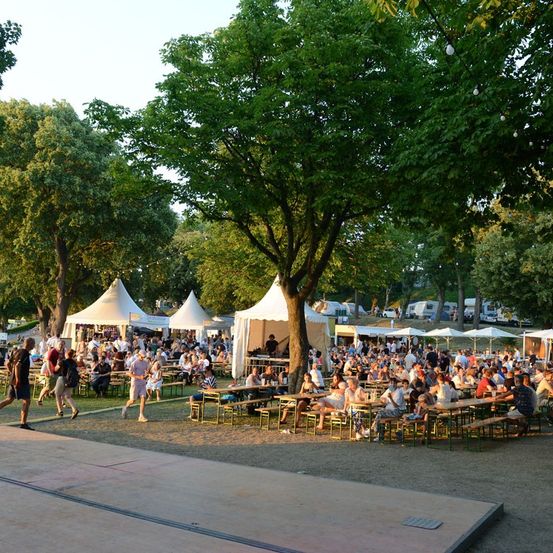 Eine große Outdoor-Veranstaltung mit vielen Menschen, die an Tischen unter weißen Zelten in einer Parkumgebung sitzen. Bäume und Grün umgeben das Gebiet, und einige Leute gehen herum.