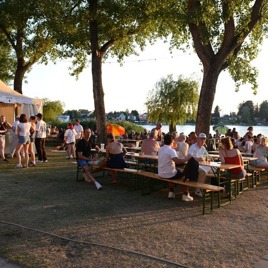 Eine Gruppe von Menschen sitzt auf Bänken um Tische in einem Park in der Nähe eines Sees, einige stehen und unterhalten sich. Bäume und ein Zelt sind im Hintergrund.